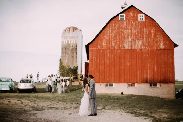 A bride and groom stand in front of a large red barn and silo on a farm, with wedding guests gathered in the background.