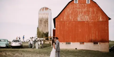 A bride and groom stand in front of a large red barn and silo on a farm, with wedding guests gathered in the background.