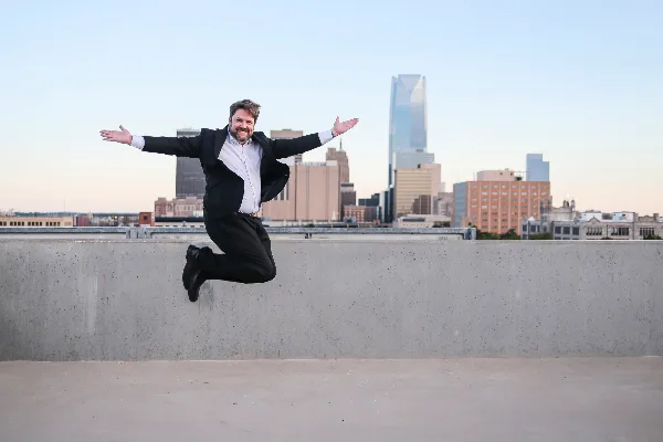 A man in a black suit jumping with arms outstretched in front of the Oklahoma City skyline.