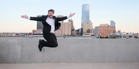 A man in a black suit jumping with arms outstretched in front of the Oklahoma City skyline.