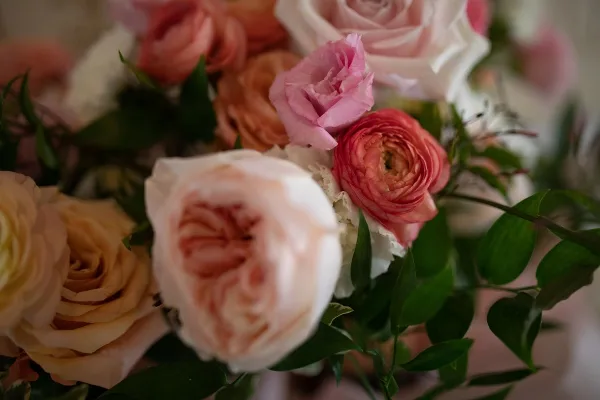 A close-up photograph of a wedding floral arrangement featuring pink and peach roses and ranunculus with green foliage.