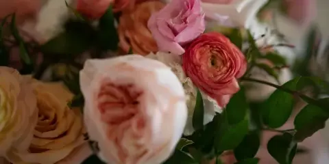 A close-up photograph of a wedding floral arrangement featuring pink and peach roses and ranunculus with green foliage.