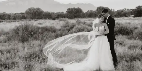 A black and white photograph of a bride and groom embracing in a grassy field with mountains in the background.