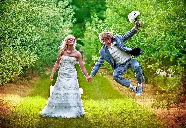 A bride in a white gown and a groom in a blue suit holding hands and jumping in a lush green garden, with the groom holding a white bouquet.
