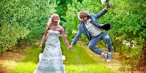 A bride in a white gown and a groom in a blue suit holding hands and jumping in a lush green garden, with the groom holding a white bouquet.