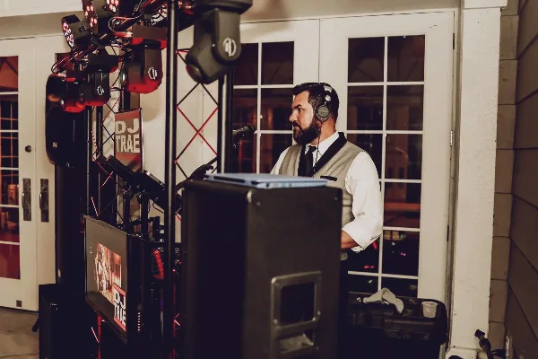 A professional DJ wearing headphones and a vest stands behind a mixing console and light rig at an event.