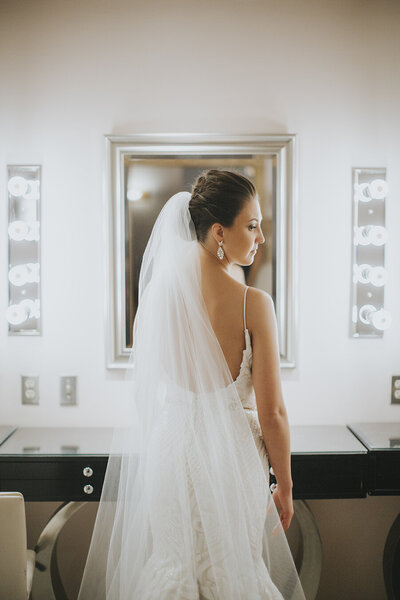 A bride in a white wedding dress and veil standing in front of a mirror with vanity lights.