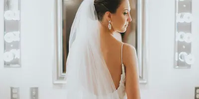 A bride in a white wedding dress and veil standing in front of a mirror with vanity lights.