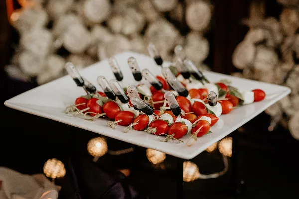 A white square plate holding an appetizer of cherry tomatoes, mozzarella, and balsamic glaze skewers, set against a blurred background of white flowers and warm lights.