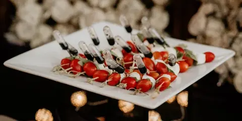 A white square plate holding an appetizer of cherry tomatoes, mozzarella, and balsamic glaze skewers, set against a blurred background of white flowers and warm lights.