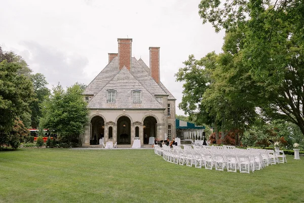 An outdoor wedding ceremony setup with rows of white chairs on a lawn in front of a historic stone building.