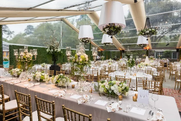 A wedding reception hall set with long tables, white linens, gold chiavari chairs, floral centerpieces, and hanging chandeliers.