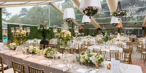 A wedding reception hall set with long tables, white linens, gold chiavari chairs, floral centerpieces, and hanging chandeliers.