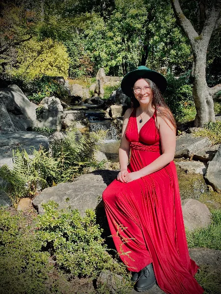 A woman in a red dress and green hat sitting on rocks near a small waterfall in a wooded area.