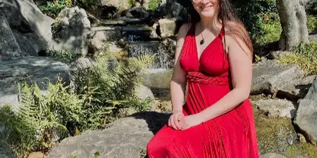 A woman in a red dress and green hat sitting on rocks near a small waterfall in a wooded area.