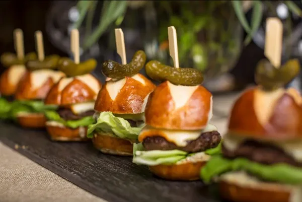 A row of mini pretzel sliders topped with cheese, lettuce, and pickles, served on a dark wooden board.
