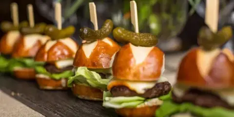 A row of mini pretzel sliders topped with cheese, lettuce, and pickles, served on a dark wooden board.