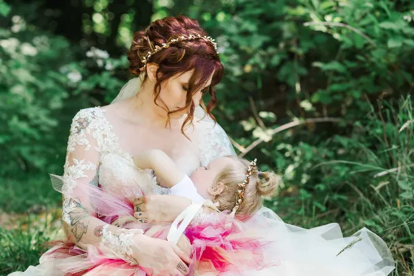 A bride in a lace wedding dress holding a young child in a pink tutu outdoors.