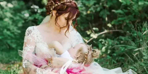 A bride in a lace wedding dress holding a young child in a pink tutu outdoors.
