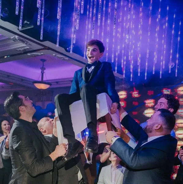 A young man in a suit is joyfully lifted on a chair by several men at a party, with colorful hanging lights in the background.
