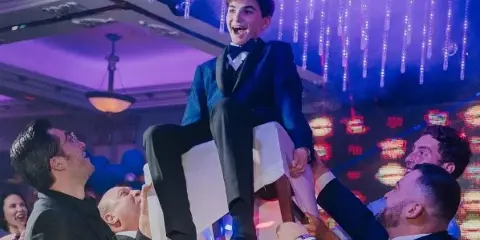 A young man in a suit is joyfully lifted on a chair by several men at a party, with colorful hanging lights in the background.