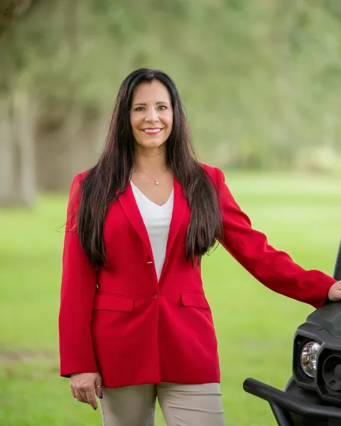 Portrait of a smiling woman with long dark hair wearing a red blazer and white top, standing outdoors in a grassy area.