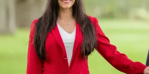 Portrait of a smiling woman with long dark hair wearing a red blazer and white top, standing outdoors in a grassy area.