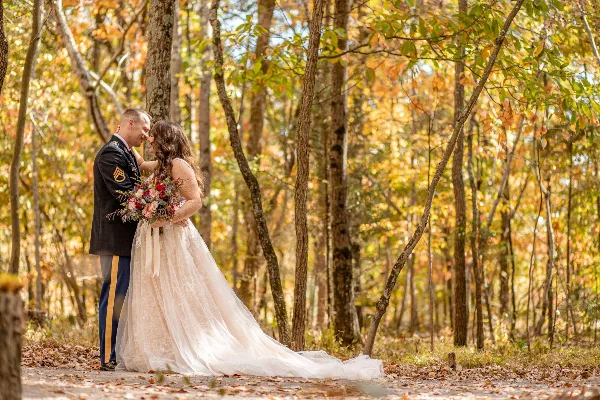 A bride in a white lace gown and a groom in a military dress uniform embrace and hold a bouquet in a sunlit autumn forest.
