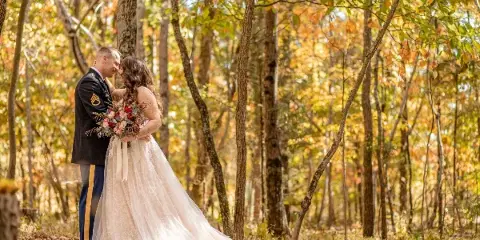 A bride in a white lace gown and a groom in a military dress uniform embrace and hold a bouquet in a sunlit autumn forest.