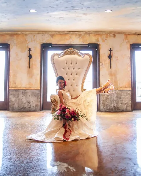 A bride in a white wedding dress sitting on a large, ornate gold and white throne chair in a venue with polished concrete floors.
