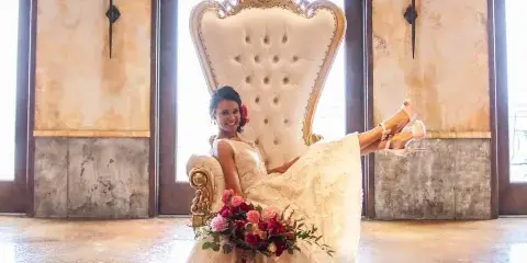 A bride in a white wedding dress sitting on a large, ornate gold and white throne chair in a venue with polished concrete floors.