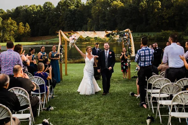 A bride and groom walking down the aisle at an outdoor wedding ceremony on a lawn near a lake.