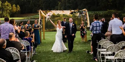 A bride and groom walking down the aisle at an outdoor wedding ceremony on a lawn near a lake.