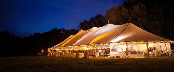 A large white event tent illuminated from within at night, set up on a grassy field with tables and chairs visible inside.