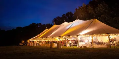 A large white event tent illuminated from within at night, set up on a grassy field with tables and chairs visible inside.