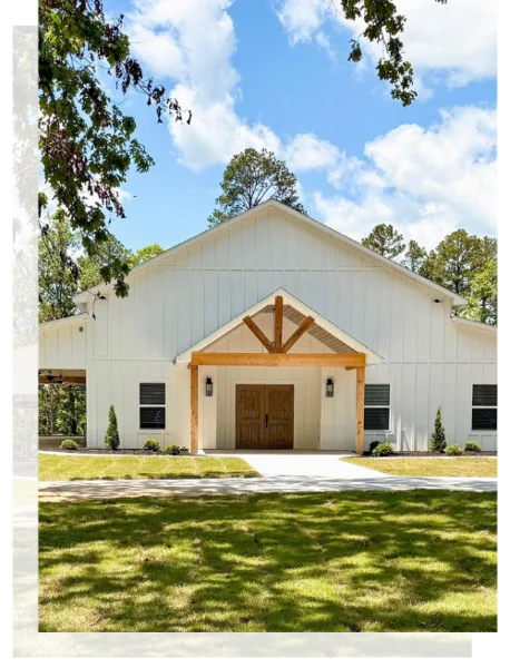Exterior view of a white, barn-style wedding venue with a wooden entrance, surrounded by green grass and trees.