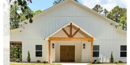 Exterior view of a white, barn-style wedding venue with a wooden entrance, surrounded by green grass and trees.