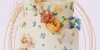A three-tiered white wedding cake decorated with colorful buttercream flowers and fresh blooms, displayed on a white pedestal cake stand.