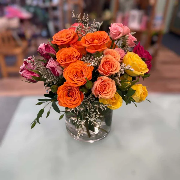 A vibrant floral arrangement consisting of orange, peach, yellow, and pink roses with white filler flowers in a clear glass vase.