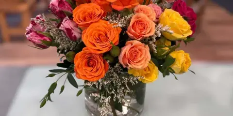 A vibrant floral arrangement consisting of orange, peach, yellow, and pink roses with white filler flowers in a clear glass vase.