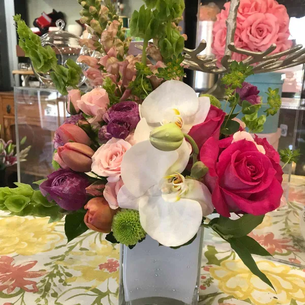 A mixed floral arrangement with pink roses, white orchids, and purple ranunculus in a square glass vase.