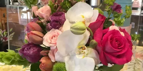 A mixed floral arrangement with pink roses, white orchids, and purple ranunculus in a square glass vase.