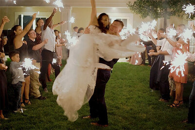 A groom lifts his bride in a white dress during a sparkler exit at a wedding reception.