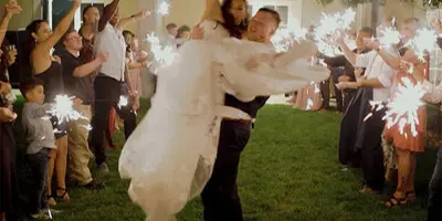 A groom lifts his bride in a white dress during a sparkler exit at a wedding reception.