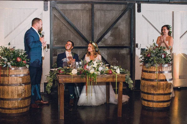 A wedding couple seated at a rustic wooden head table decorated with lush floral arrangements and greenery, flanked by wine barrels with floral displays.