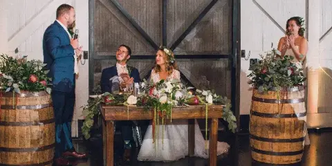 A wedding couple seated at a rustic wooden head table decorated with lush floral arrangements and greenery, flanked by wine barrels with floral displays.