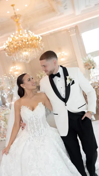 A bride and groom posing together in a luxurious ballroom with a large chandelier.