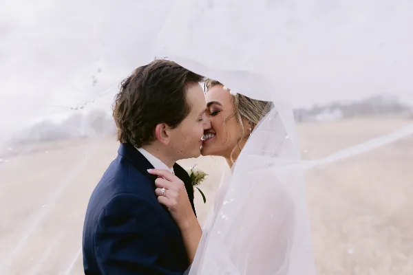 A bride and groom sharing a kiss under a veil in a field.