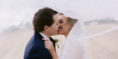 A bride and groom sharing a kiss under a veil in a field.