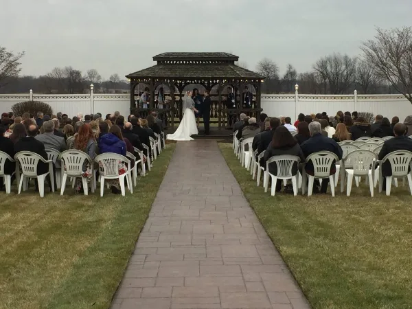 Outdoor wedding ceremony at a gazebo with a paved aisle and seated guests in a landscaped courtyard.